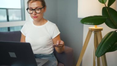 Gözlüklü bir kadın rahat bir odada kanepeye oturur ve kredi kartı ve akıllı telefon kullanarak online alışveriş yapar. İnternetten alışveriş, yaşam tarzı teknolojisi.
