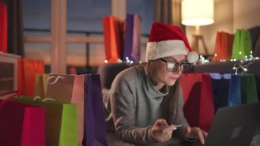 Noel Baba şapkası takan mutlu kadın halıda yatıyor ve kredi kartı ve dizüstü bilgisayar kullanarak online alışveriş yapıyor. Alışveriş torbaları..