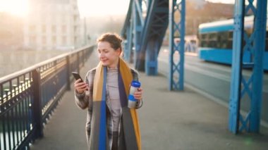 Paltolu beyaz bir iş kadınının portresi, soğuk bir sabahta köprüde yürüyor, kahve içiyor ve akıllı telefon kullanıyor. İletişim, iş günü, yoğun yaşam konsepti. Yavaş çekim