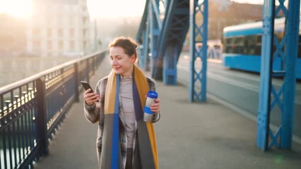 Portret van een jonge blanke zakenvrouw in een jas, die op een ijzige ochtend over de brug loopt, koffie drinkt en een smartphone gebruikt. Communicatie, werkdag, druk levensconcept. Langzame beweging — Stockvideo