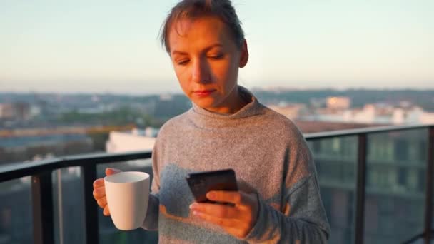 Mulher começa seu dia com uma xícara de chá ou café e verificar e-mails em seu smartphone na varanda ao amanhecer. — Vídeo de Stock