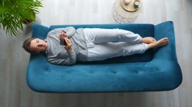 Akıllı telefon kullanan, cep telefonu kullanan, komik videolar izleyen, sosyal medyada sohbet eden, evdeki kanepede uzanan mutlu ve rahat bir kadının genel görüntüsü..