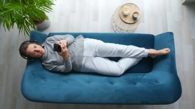Kablosuz kulaklıklı kadın müzik dinlerken bir yandan bir yana bir yana hareket ediyor ve mobil uygulamalar kullanıyor ya da evdeki kanepede uzanan akıllı telefondan sosyal ağlarda iletişim kuruyor.