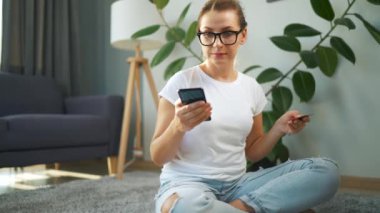 Gözlüklü kadın yerde oturuyor ve kredi kartı ve akıllı telefon kullanarak online alışveriş yapıyor. İnternetten alışveriş, yaşam tarzı teknolojisi.