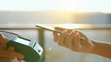 Yakın plan, kadın banka terminalindeki siparişi ödemek için akıllı telefon nakitsiz cüzdan teknolojisi kullanıyor. Kart makinesi erkek eliyle güneş ışığı arkaplanında