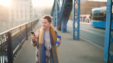 Paltolu beyaz bir iş kadınının portresi, soğuk bir sabahta köprüde yürüyor, kahve içiyor ve akıllı telefon kullanıyor. İletişim, iş günü, yoğun yaşam konsepti.