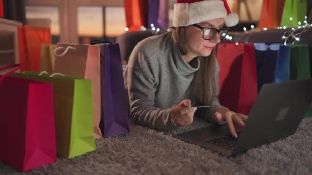 Happy woman with glasses wearing a santa claus hat is lying on the carpet and makes an online purchase using a credit card and laptop. Shopping bags around. — Stock Video