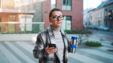 Beyaz tenli, gözlüklü ve ceketli genç bir iş kadınının portresi iş bölgesinde termik bardakla yürüyor ve akıllı telefon kullanıyor. İletişim, iş günü, yoğun yaşam konsepti.