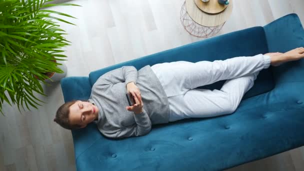 Накладний знімок щасливої розслабленої жінки, що тримає смартфон, використовує мобільні додатки, дивиться смішне відео, весело спілкується в соціальних мережах, лежить на дивані вдома. Камера обертається проти годинникової стрілки Ліцензійні Стокові Відеоролики