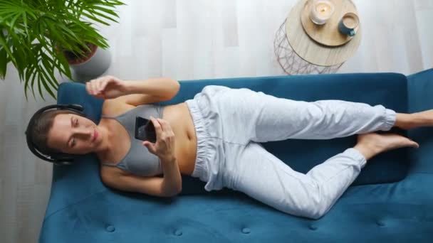 Жінка в бездротових навушниках слухає музику і використовує мобільні додатки або спілкується в соціальних мережах на смартфоні, лежачи на дивані вдома, вид зверху. Камера обертається за годинниковою стрілкою Стокове Відео 