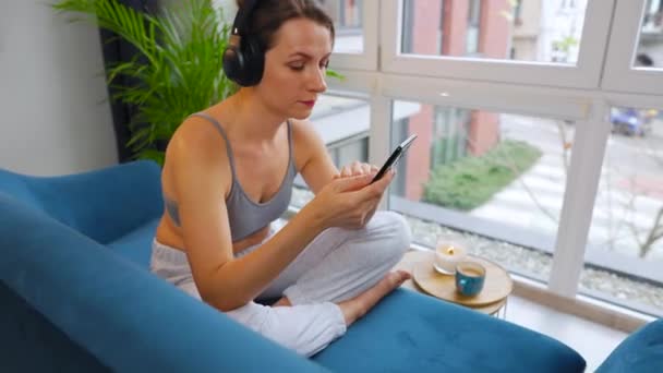 Kablosuz kulaklık takan, müzik dinleyen ve cep telefonu uygulamaları kullanan ya da akıllı telefonlu bir kadın evdeki kanepede oturuyor. — Stok video
