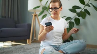 Gözlüklü kadın yerde oturuyor ve kredi kartı ve akıllı telefon kullanarak online alışveriş yapıyor. İnternetten alışveriş, yaşam tarzı teknolojisi.