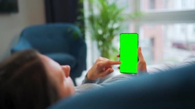 Kadın evde kanepede yatıyor ve dikey modda yeşil maket ekranlı akıllı telefon kullanıyor. Kız internette geziniyor, içeriği, videoları, blogları izliyor.