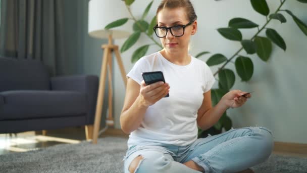Mujer con gafas se sienta en el suelo y hace una compra en línea con una tarjeta de crédito y un teléfono inteligente. Compras en línea, tecnología de estilo de vida — Vídeos de Stock