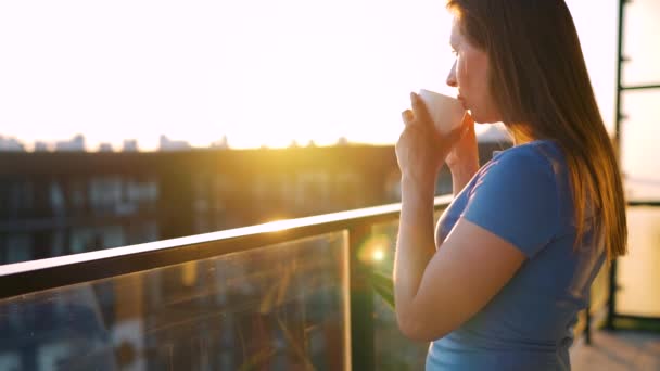 Mulher com uma xícara de café em pé na varanda e admirar o pôr do sol. Movimento lento — Vídeo de Stock