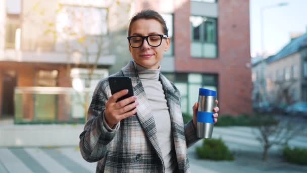 Portrait of a young caucasian businesswoman with glasses and a coat walks through the business district, with thermo cup and using smartphone. Communication, work day, busy life concept. Slow motion — Stock Video