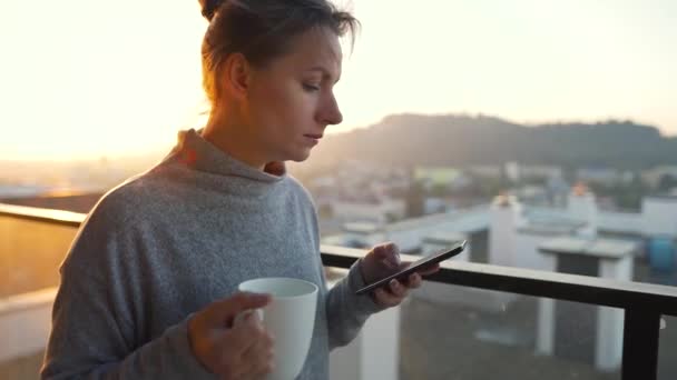 La mujer comienza el día con una taza de té o café y revisando los correos electrónicos en su teléfono inteligente en el balcón al amanecer.. — Vídeo de stock