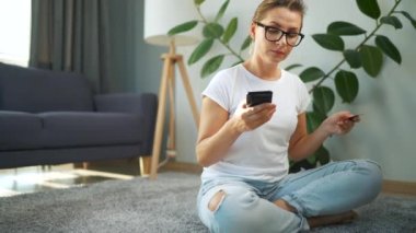 Gözlüklü kadın yerde oturuyor ve kredi kartı ve akıllı telefon kullanarak online alışveriş yapıyor. İnternetten alışveriş, yaşam tarzı teknolojisi.
