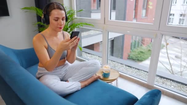 Kablosuz kulaklık takan, müzik dinleyen ve cep telefonu uygulamaları kullanan ya da akıllı telefonlu bir kadın evdeki kanepede oturuyor. — Stok video