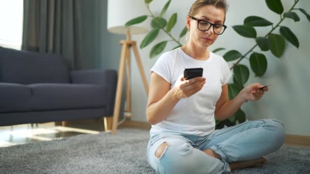 Frau mit Brille sitzt auf dem Boden und kauft online mit Kreditkarte und Smartphone ein. Online-Shopping, Lifestyletechnologie — Stockvideo