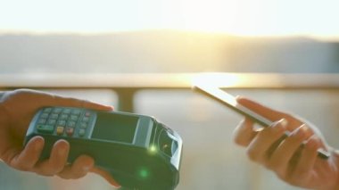 Yakın plan, kadın banka terminalindeki siparişi ödemek için akıllı telefon nakitsiz cüzdan teknolojisi kullanıyor. Kart makinesi erkek eliyle güneş ışığı arkaplanında