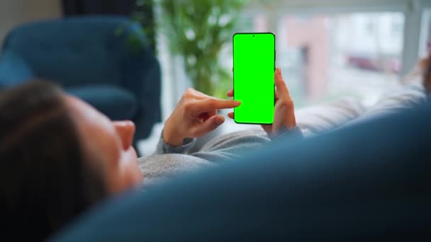 Mujer se encuentra en casa en el sofá y el uso de teléfono inteligente con pantalla de maqueta verde en modo vertical. Chica navegando por Internet, viendo contenido, videos, blogs. — Vídeo de stock
