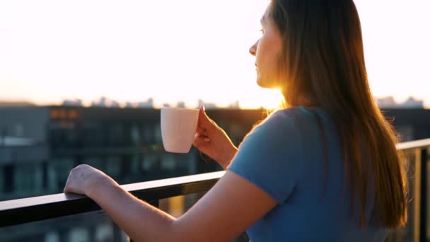 Elinde bir fincan kahve olan kadın balkonda duruyor ve gün batımını seyrediyor. Yavaş çekim — Stok video