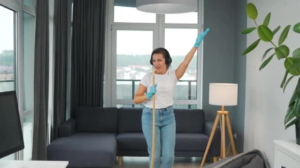 Frau mit Kopfhörer putzt das Haus und hat Spaß beim Singen mit dem Besen wie ein Star bei einem Konzert. Zeitlupe — Stockvideo