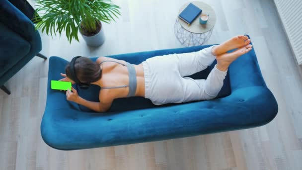 Женщина в беспроводных наушниках, слушающая музыку и использующая мобильные приложения или общающаяся в социальных сетях на смартфоне с зеленым макетом экрана, лежащая на диване дома, вид сверху — стоковое видео