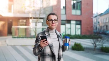 Beyaz tenli, gözlüklü ve ceketli genç bir iş kadınının portresi iş bölgesinde termik bardakla yürüyor ve akıllı telefon kullanıyor. İletişim, iş günü, yoğun yaşam konsepti