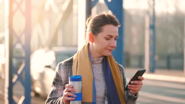 Portrait of a young caucasian businesswoman in a coat, walking around the city on a frosty morning, drinking coffee and using smartphone. Communication, work day, busy life concept. — Video Stock