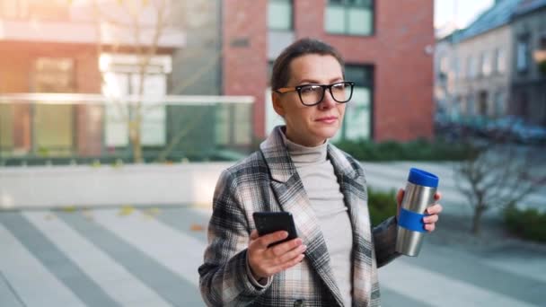 Ritratto di una giovane donna d'affari caucasica con occhiali e cappotto passeggia per il quartiere degli affari, con thermo cup e utilizzando lo smartphone. Comunicazione, giornata di lavoro, concetto di vita frenetica. Rallentatore — Video Stock