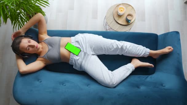 Felülről lövés nyugodt nő bekapcsolja a zenét egy alkalmazás egy okostelefon zöld modell képernyőn, és hallgatja, hogy a fejhallgató, feküdt a kanapén otthon. — Stock videók