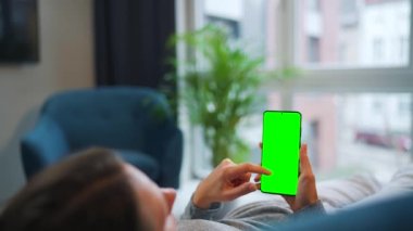 Kadın evde kanepede yatıyor ve dikey modda yeşil maket ekranlı akıllı telefon kullanıyor. Kız internette geziniyor, içeriği, videoları, blogları izliyor.
