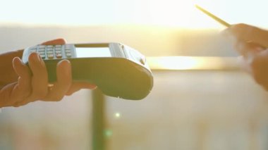 Yakın plan, kadın banka terminalindeki siparişi ödemek için akıllı telefon nakitsiz cüzdan teknolojisi kullanıyor. Kart makinesi erkek eliyle güneş ışığı arkaplanında