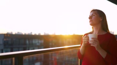 Kırmızı elbiseli bir kadın elinde bir fincan kahveyle balkonda duruyor ve gün batımını seyrediyor. Yavaş çekim