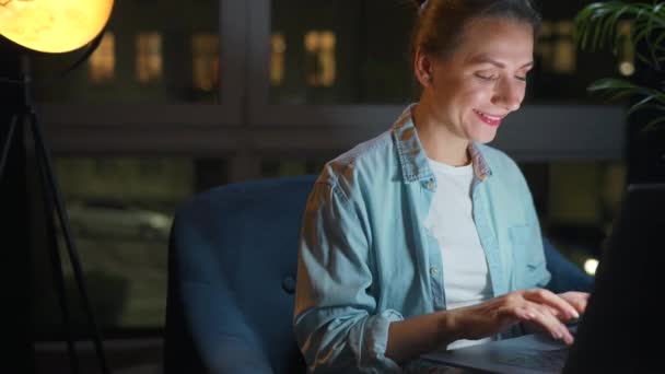 Donna sorridente è seduto sulla poltrona e lavorare su un computer portatile o chattare con qualcuno di notte. Concetto di lavoro a distanza. — Video Stock