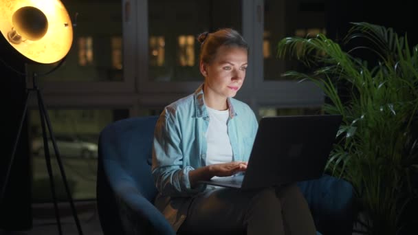 La mujer está sentada en el sillón y trabajando en un portátil por la noche. Concepto de trabajo remoto. — Vídeos de Stock