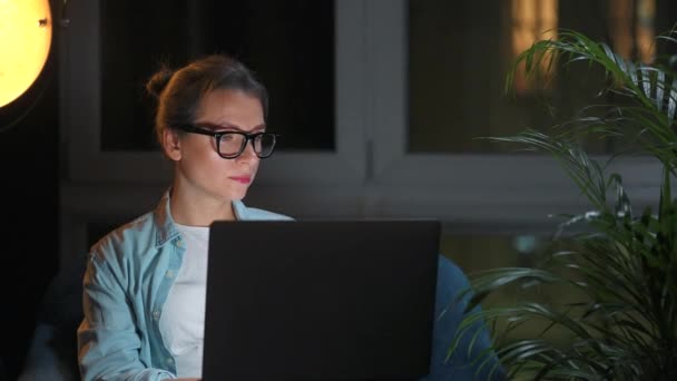 Mujer con gafas está sentado en el sillón y trabajando en un ordenador portátil por la noche. Concepto de trabajo remoto. — Vídeos de Stock