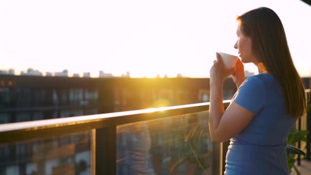 Mujer con una taza de café de pie en el balcón y admirar la puesta de sol. Movimiento lento — Vídeos de Stock