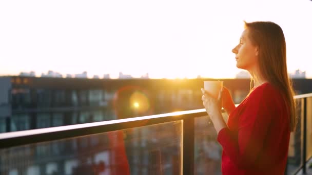 Kırmızı elbiseli kadın, elinde bir fincan kahveyle balkonda duruyor ve gün batımını seyrediyor.. — Stok video