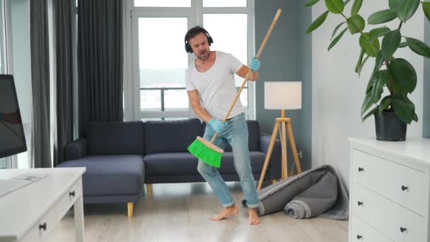 Homem ouve música com fones de ouvido e limpar a casa e se divertir dançando com uma vassoura. Movimento lento — Vídeo de Stock