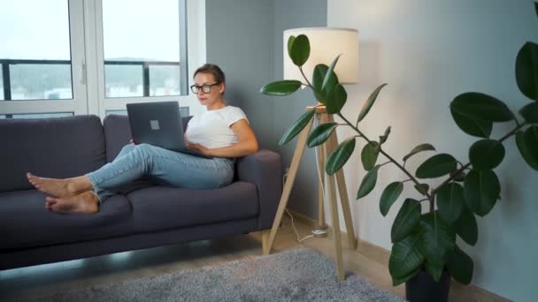 Kobieta w okularach siedzi na kanapie i pracuje na laptopie. Pojęcie pracy zdalnej. — Wideo stockowe