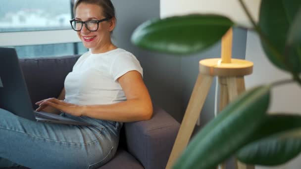 Uśmiechnięta kobieta w okularach siedzi na kanapie i pracuje nad laptopem albo z kimś rozmawia. Pojęcie pracy zdalnej. — Wideo stockowe