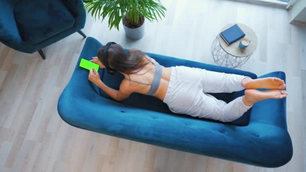 Donna con cuffie wireless che ascolta musica e utilizza app mobili o comunica sui social network su smartphone con schermo modello verde, sdraiata sul divano di casa, vista dall'alto — Video Stock