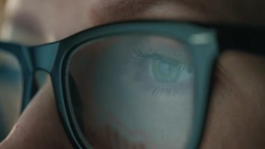 Gözlüklü kadın monitöre bakıyor ve çizelgeler ve analizlerle çalışıyor. Monitör ekranı gözlüklere yansıyor. Gece çalış. Aşırı yakın çekim