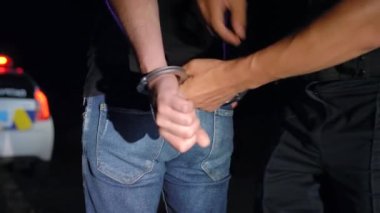 Polis memuru gece tutuklanan adama kelepçe takıyor ve onu polis arabasına götürüyor.