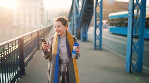 Retrato de uma jovem mulher de negócios caucasiana de casaco, atravessando a ponte em uma manhã gelada, tomando café e usando smartphone. Comunicação, dia de trabalho, conceito de vida ocupada. Movimento lento — Vídeo de Stock