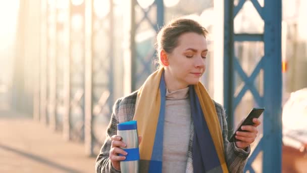 Retrato de una joven mujer de negocios caucásica en un abrigo, caminando por la ciudad en una mañana helada, tomando café y usando un teléfono inteligente. Comunicación, día de trabajo, concepto de vida ocupada. Movimiento lento — Vídeo de stock