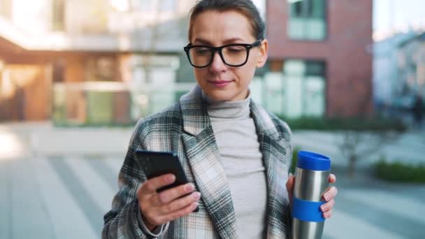 Portrait of a young caucasian businesswoman with glasses and a coat walks through the business district, with thermo cup and using smartphone. Communication, work day, busy life concept. Slow motion — Stockvideo